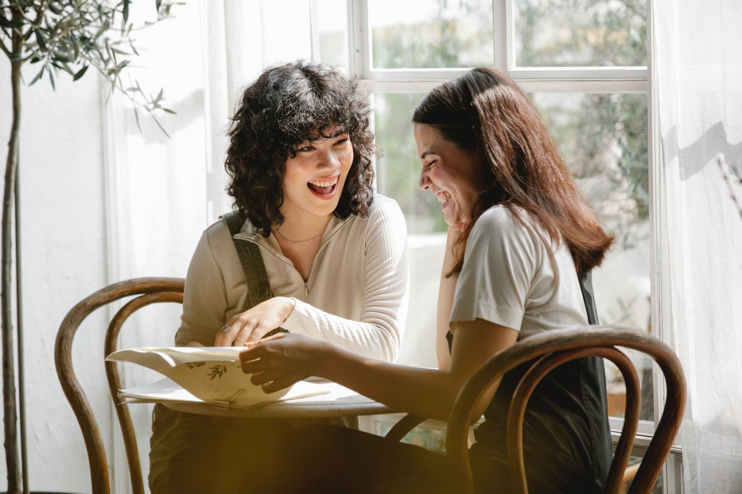 Two women sitting at a table, reading, and laughing